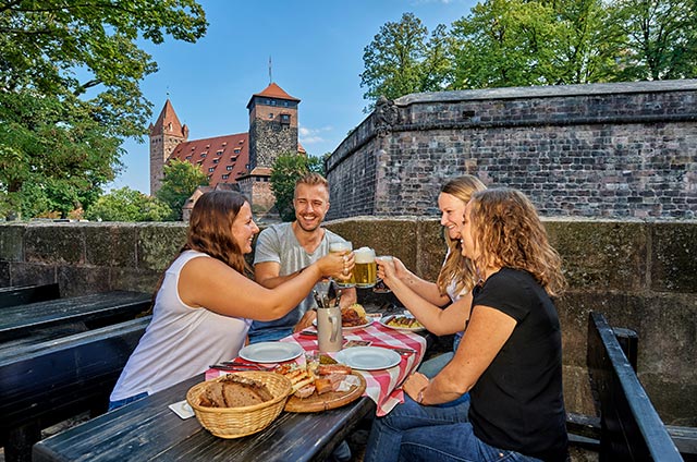 Vier Personen sitzen bei der Brotzeit vor einer Burgmauer und stoßen mit Bierkrügen an